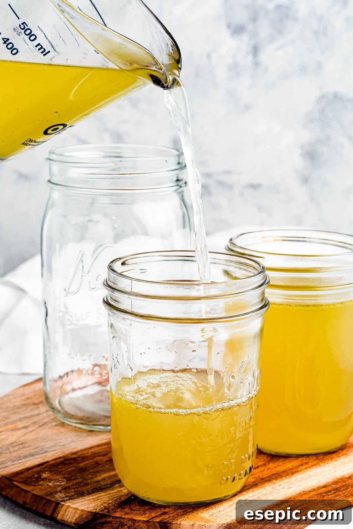 Homemade chicken broth being poured from a large clear measuring cup into several glass jars, highlighting the beautiful golden color and freshness.