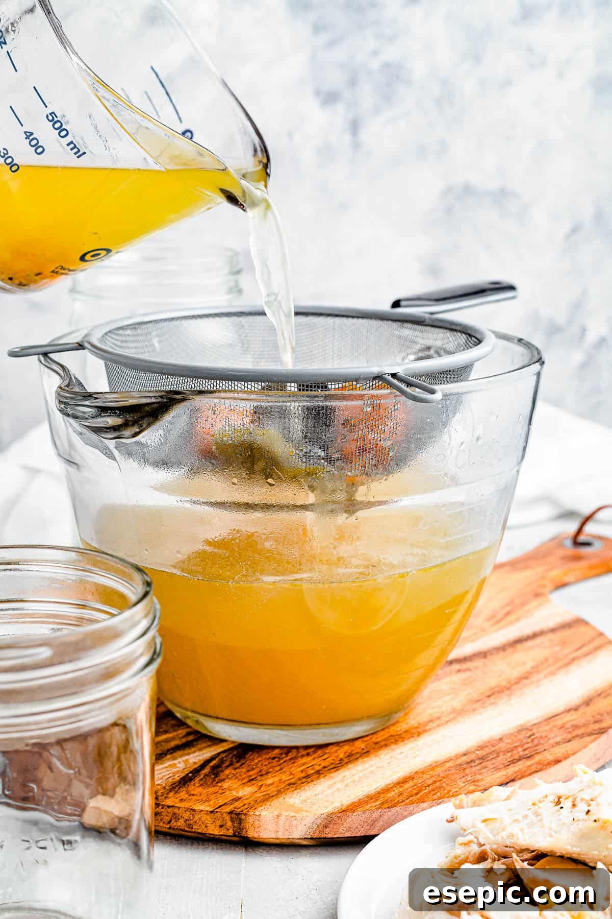 Golden chicken broth being carefully poured from a large measuring cup through a fine-mesh sieve into a mixing bowl, illustrating the straining process.