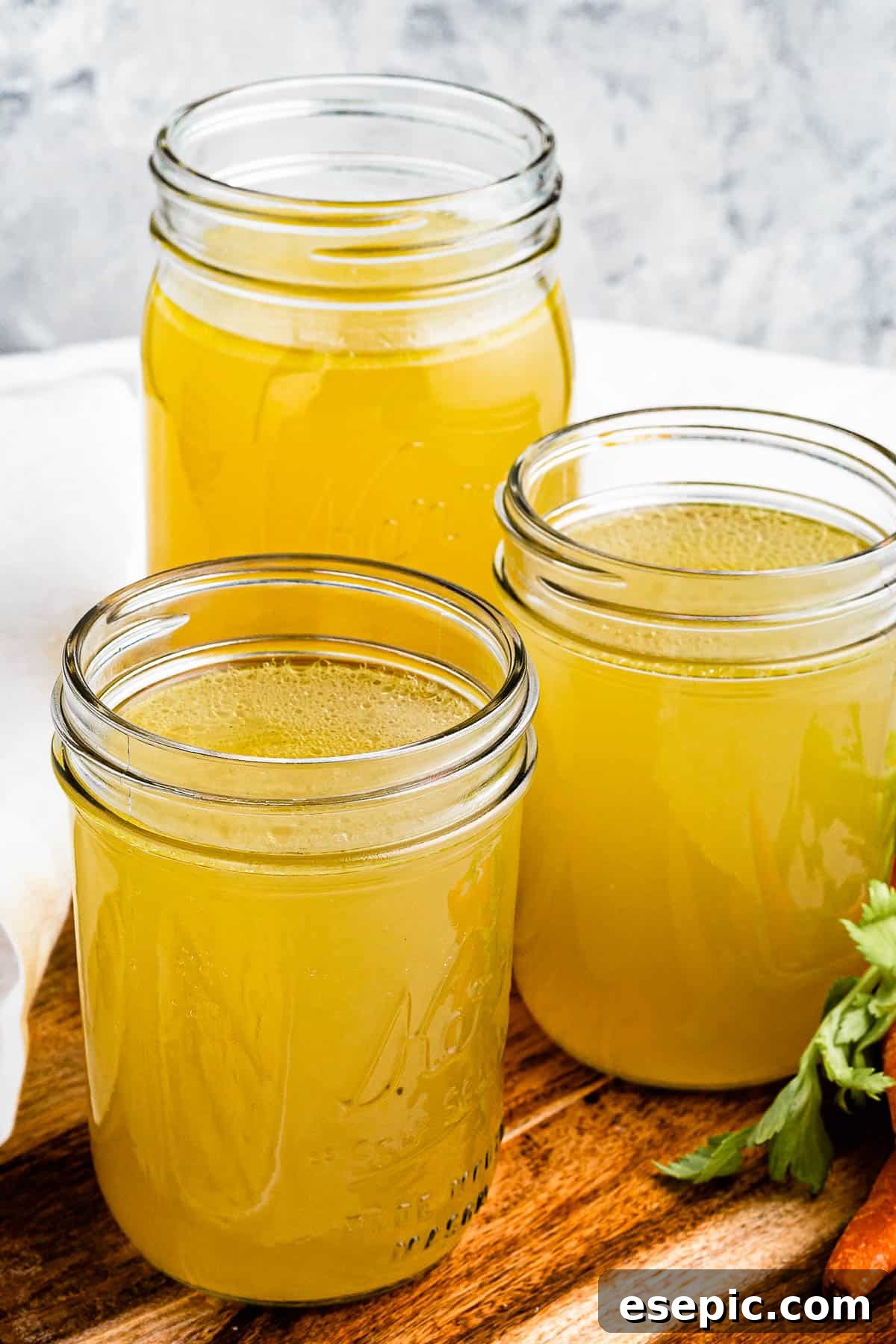 Three jars of golden homemade chicken broth resting on a wooden cutting board, with fresh raw carrots adding a touch of natural beauty to the scene.