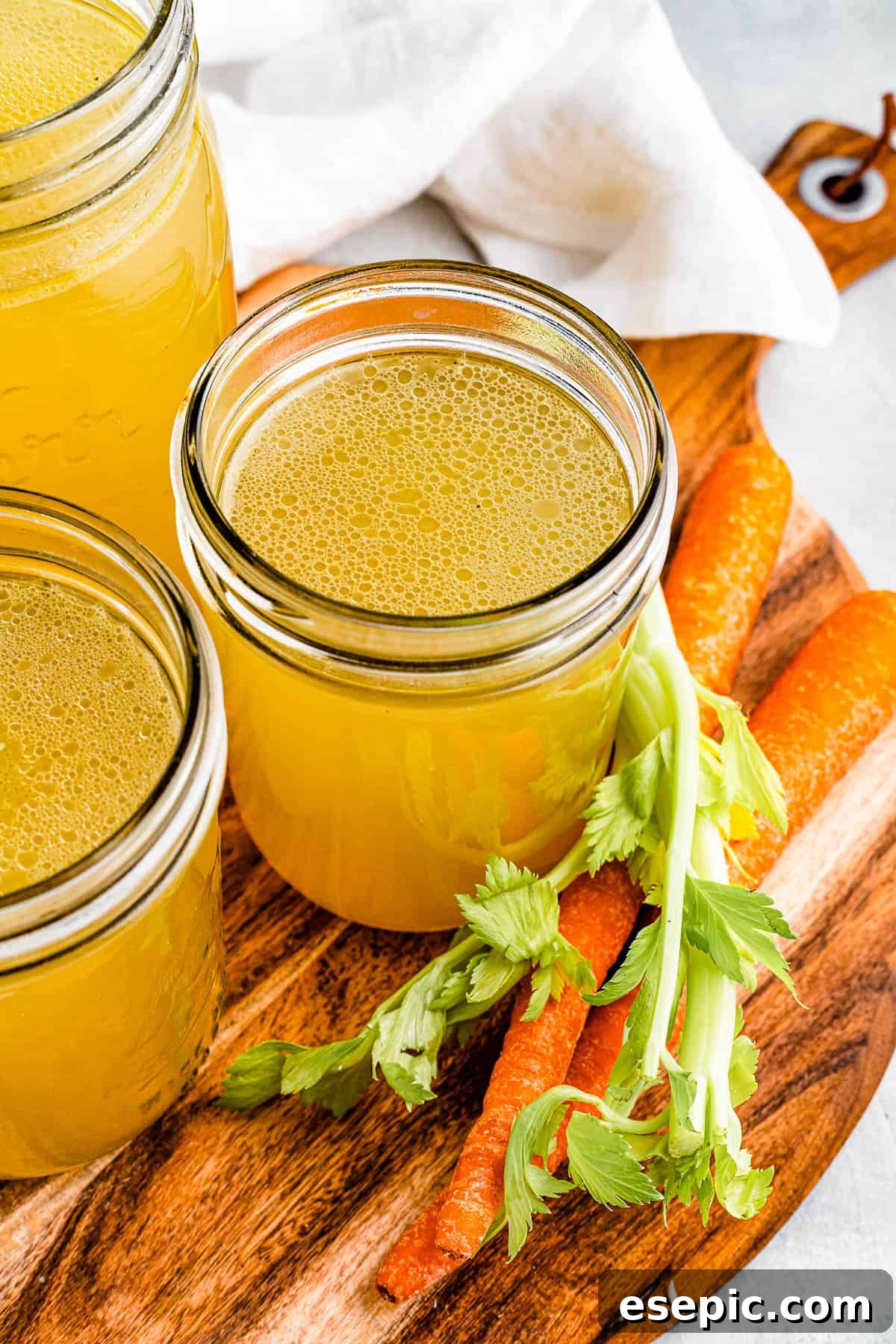 Three glass jars of freshly made chicken broth neatly arranged on a wooden cutting board, with whole raw carrots beside them, ready for culinary use.