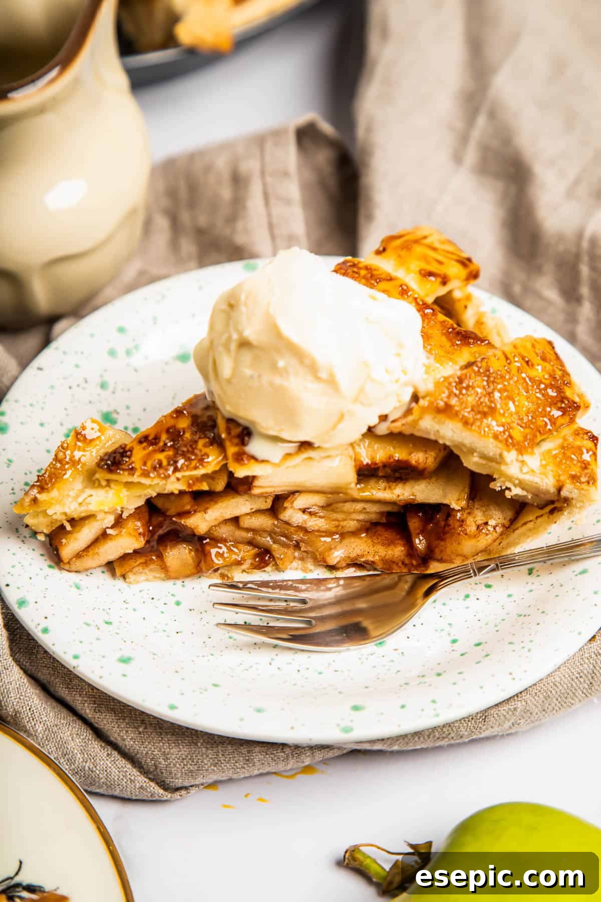 This classic yet easy and delicious Apple Pie recipe is made from scratch, with a buttery, flaky pie crust, fresh apple slices, pie spices, and brown sugar. A perfectly baked slice of homemade apple pie, topped with a scoop of vanilla ice cream, served on a white plate with a fork.