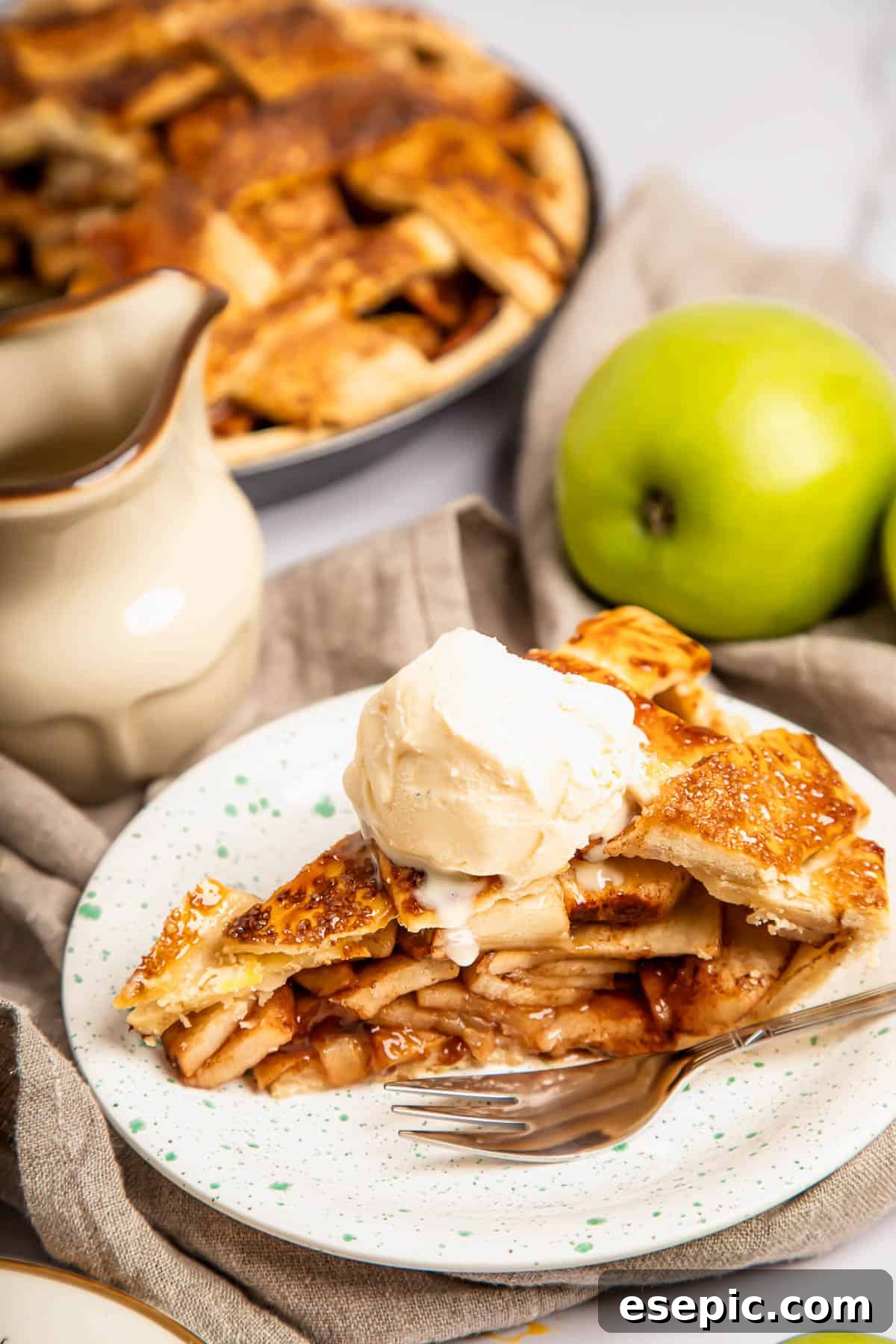 This classic yet easy and delicious Apple Pie recipe is made from scratch, with a buttery, flaky pie crust, fresh apple slices, pie spices, and brown sugar. A delectable slice of homemade apple pie crowned with a generous scoop of vanilla ice cream, presented on a white plate with a fork nearby.