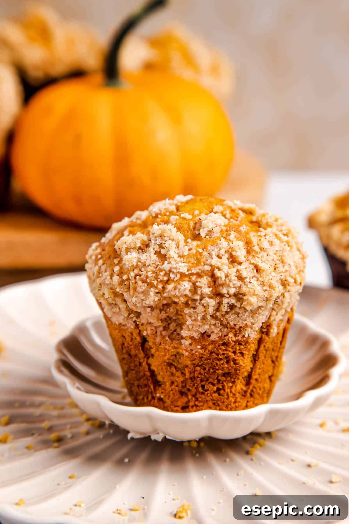 A single, perfectly baked pumpkin muffin adorned with a golden cinnamon crumble sits on a white plate, with a soft-focus background of more muffins.