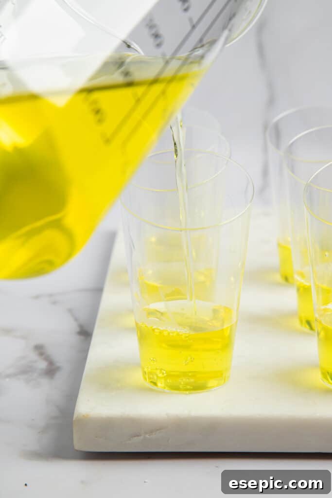 Bright yellow lemon jello being carefully poured into the bottom of several shot glasses.