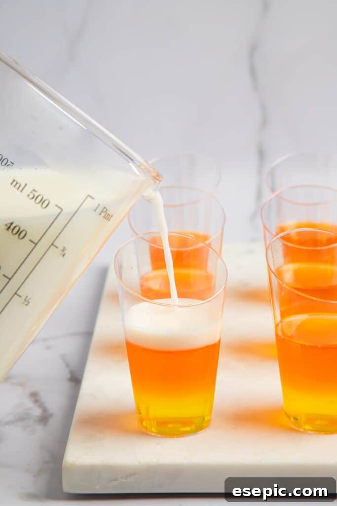 A creamy white jello mixture being gently poured onto the orange layer in jello shot glasses, completing the candy corn look.