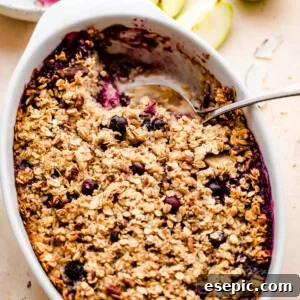 Warm Apple Blueberry Oatmeal Bake 7 overhead shot of baked oatmeal in a white oval baking dish with a green apple set on the side, and a plate with oatmeal right above it.