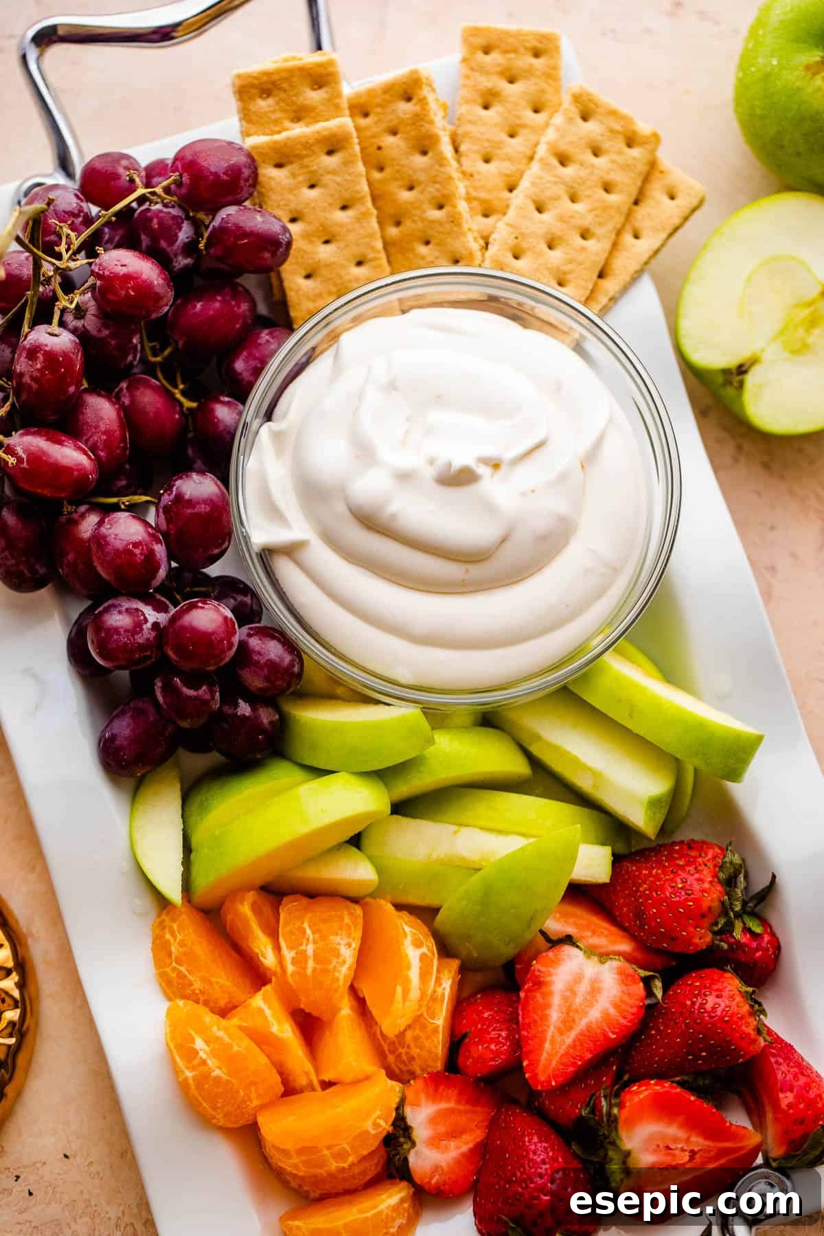 A vibrant serving platter featuring a bowl of creamy coconut fruit dip, neatly arranged with slices of green apple, juicy tangerine segments, fresh strawberries, plump grapes, and crispy graham crackers.