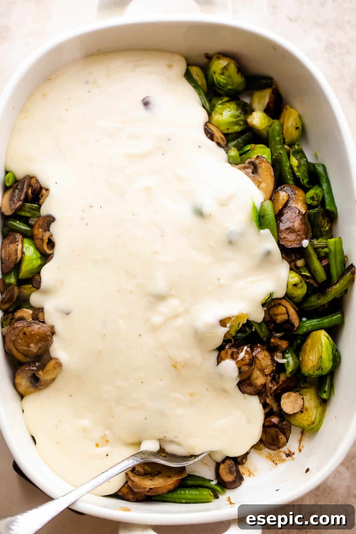 Pouring rich, creamy Parmesan sauce over sautéed green beans, Brussels sprouts, and mushrooms in a white baking dish.