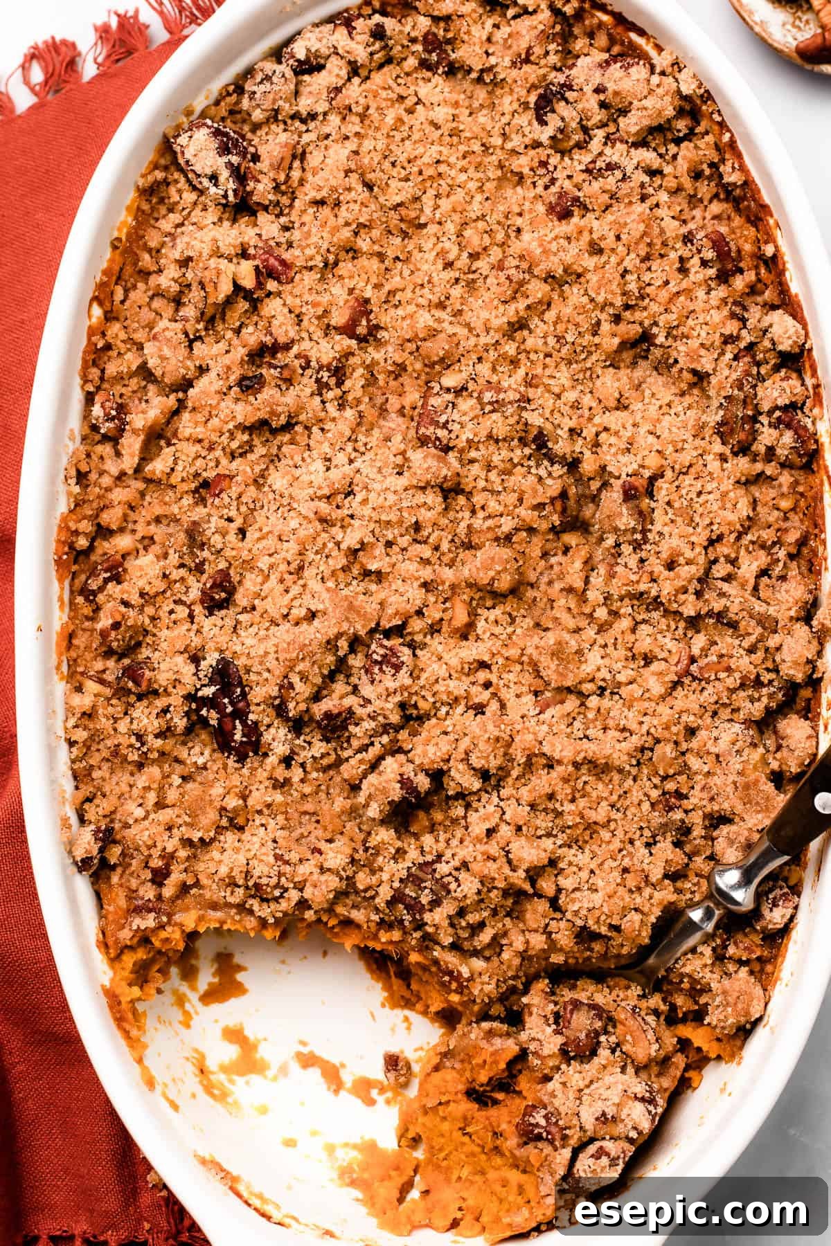 A baking dish of sweet potatoes and crumb topping, on a marble background. A serving of the casserole has been scooped out, and a spoon is resting in the dish.