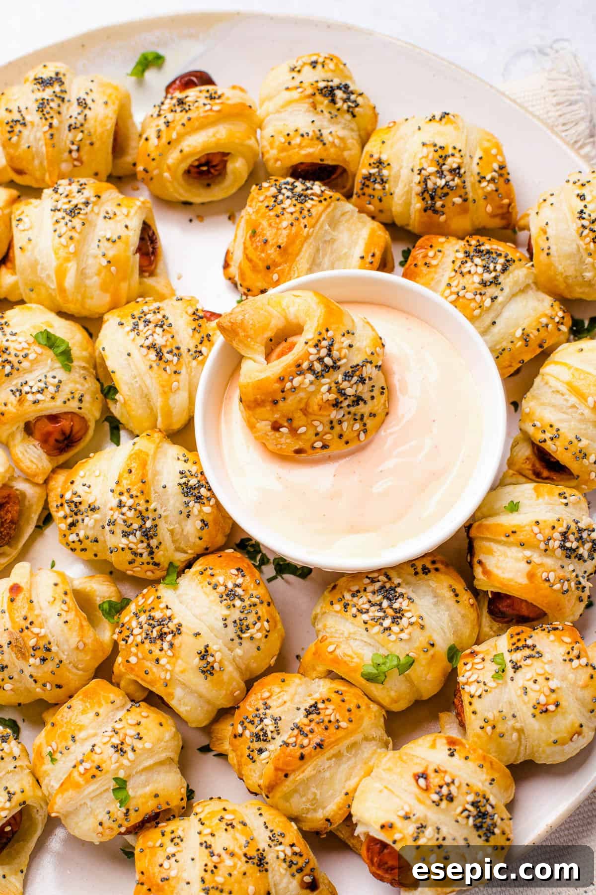 Super easy to make homemade Pigs in a Blanket are a delicious snack prepared with savory mini-sausages wrapped in golden, flaky puff pastry. A platter of pigs in a blanket with a bowl of dipping sauce in the center. One pastry is dipped in the sauce.