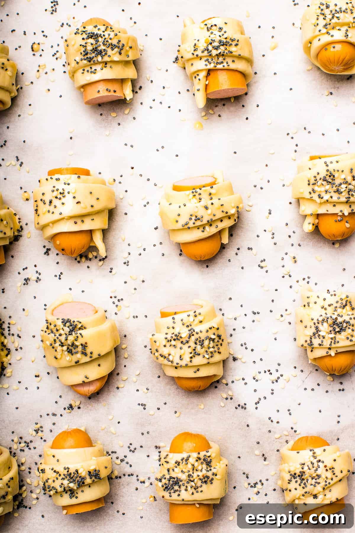 Unbaked pigs in a blanket with egg wash and seeds. Unbaked pastries brushed with egg wash and sprinkled with sesame seeds and poppy seeds.