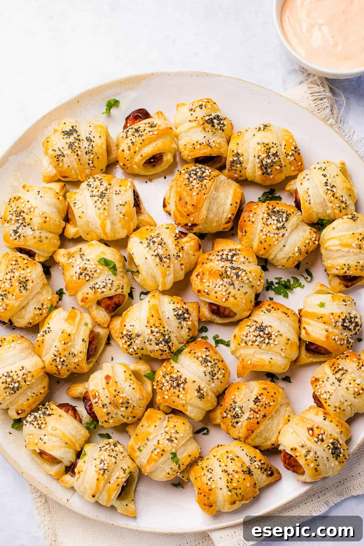 Platter of golden pigs in a blanket with dipping sauce. A serving platter holding about two dozen pigs in a blanket. A bowl of dipping sauce sits nearby.
