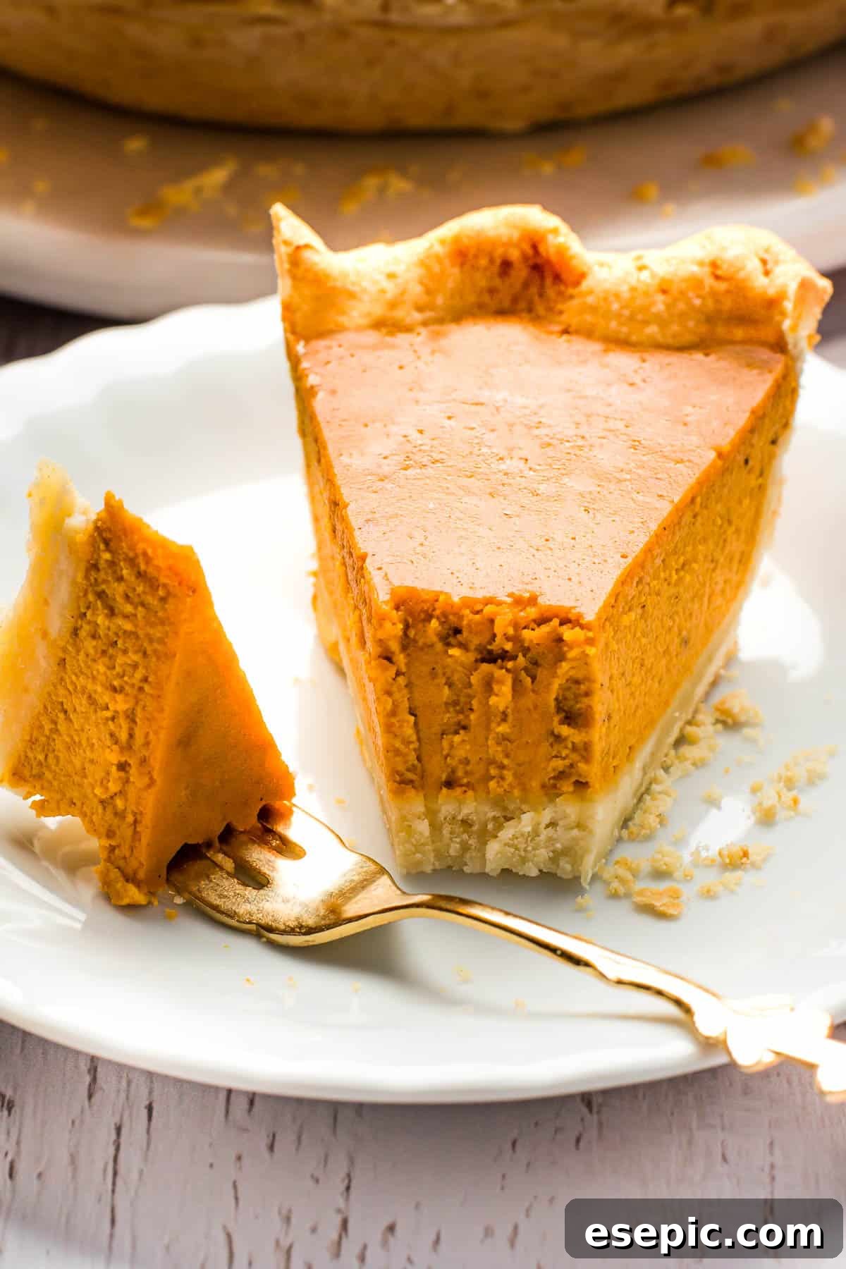 This easy homemade Pumpkin Pie recipe, featuring a tender crust, warm spices, and dulce de leche pumpkin filling, is a perfect holiday favorite that everyone will adore. A close-up shot of a single slice of homemade pumpkin pie, showcasing its smooth, creamy filling and golden, crimped crust, elegantly presented on a white plate with a silver fork.