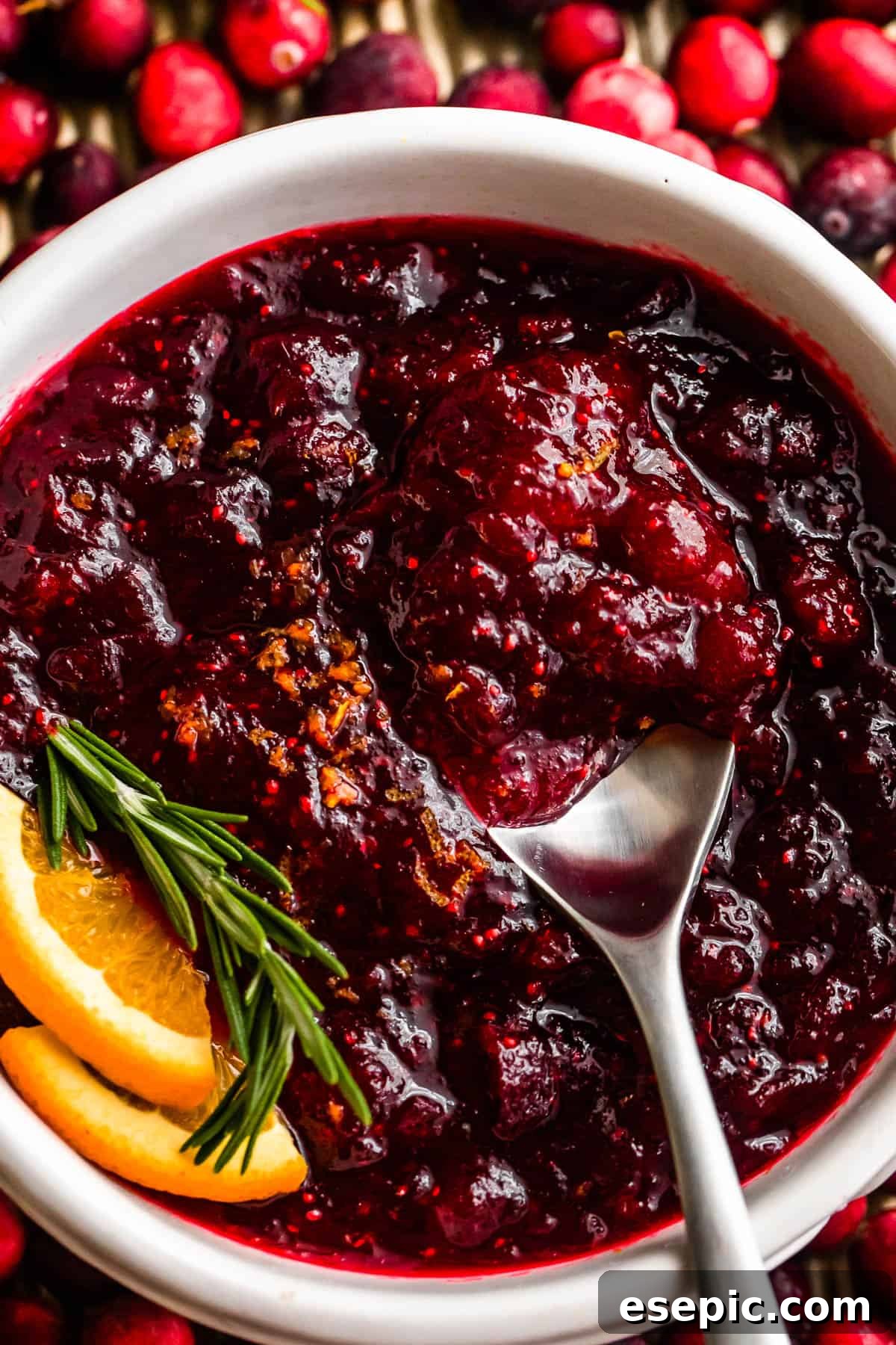 overhead close up shot of a spoon inside of a bowl filled with cranberry sauce and garnished with rosemary sprig plus orange slices