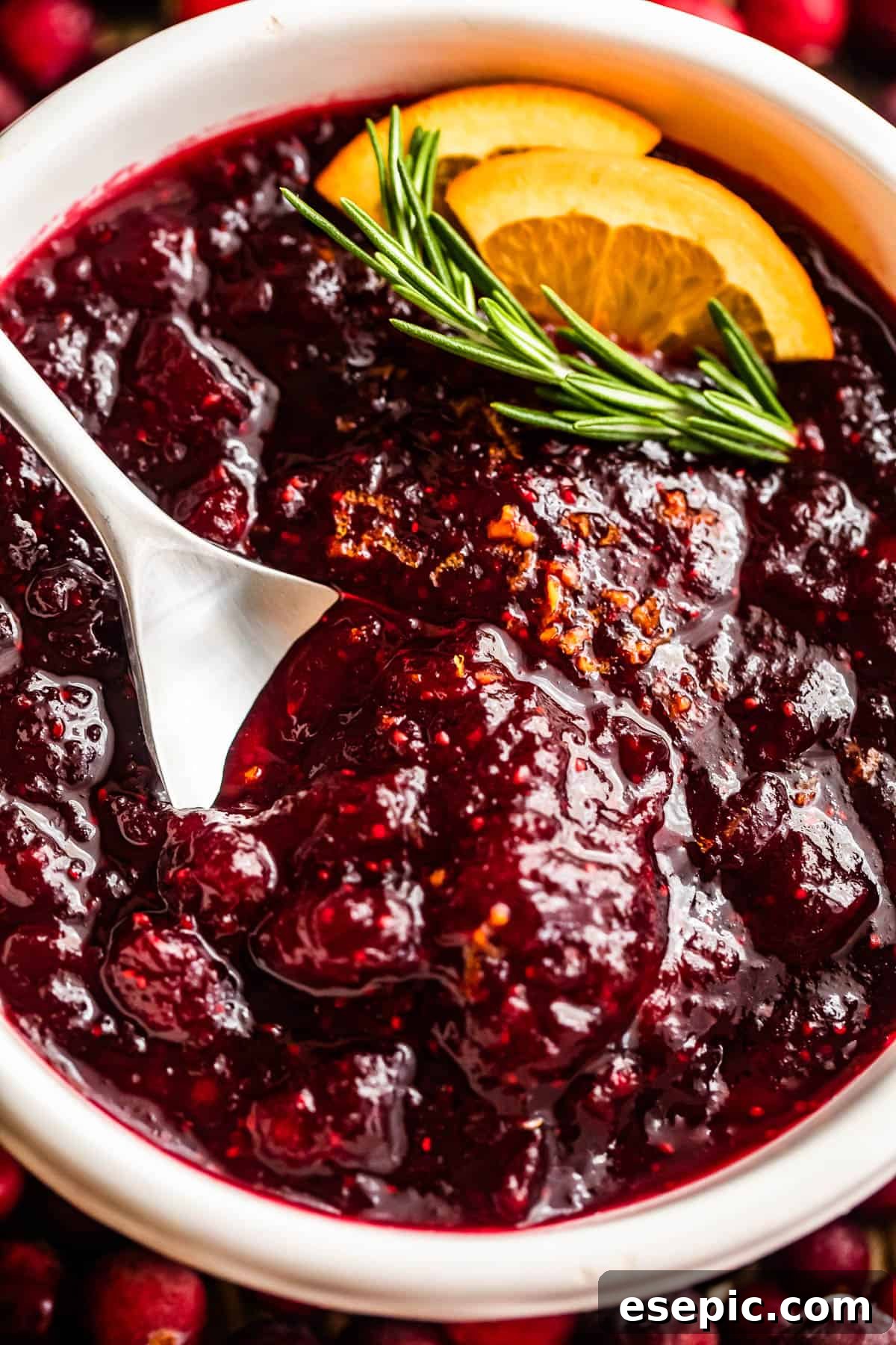 overhead close up shot of a spoon inside of a bowl filled with cranberry sauce and garnished with rosemary sprig plus orange slices